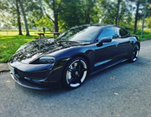 Porsche Taycan noire stationnée sur une route bordée d’arbres, véhicule électrique haut de gamme convoyée par KirriGo.
